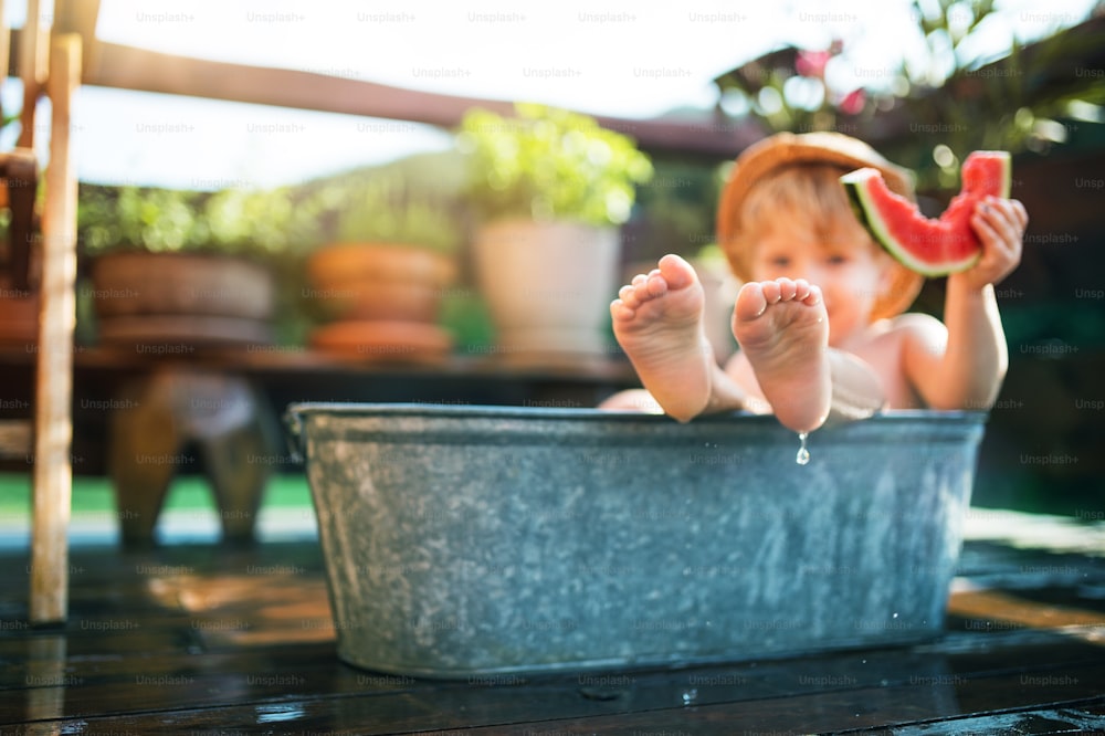 Niño pequeño feliz con un sombrero en la bañera al aire libre en el jardín en verano, comiendo sandía.
