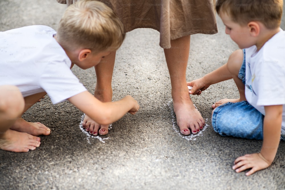 Zwei kleine Söhne, die an einem Sommertag mit einer nicht wiederzuerkennenden Mutter auf einer Straße im Park spielen und mit Kreide Umrisse ihrer Füße zeichnen.