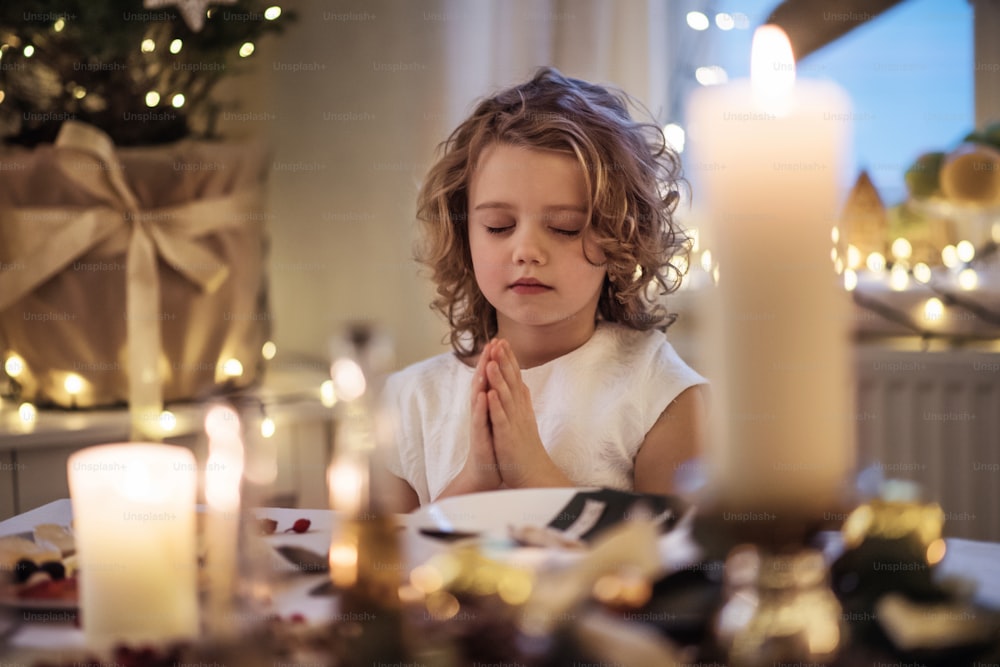 Retrato da menina pequena sentada à mesa dentro de casa no Natal, orando.