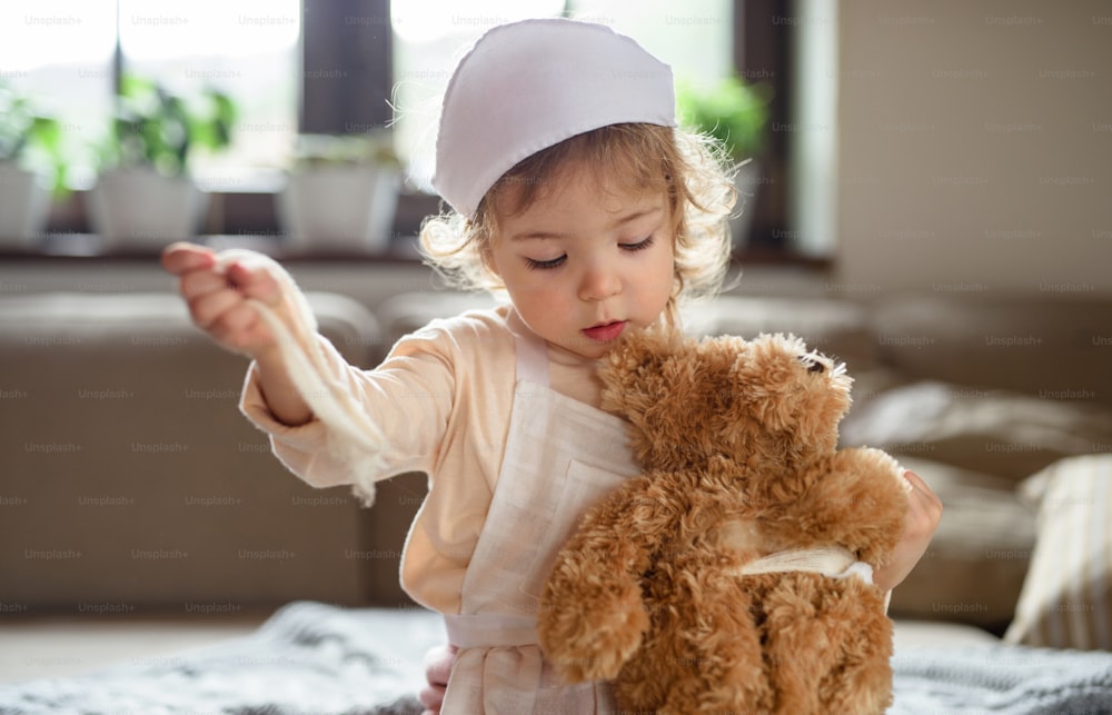 Small toddler girl with doctor uniform indoors at home, playing with teddy bear.