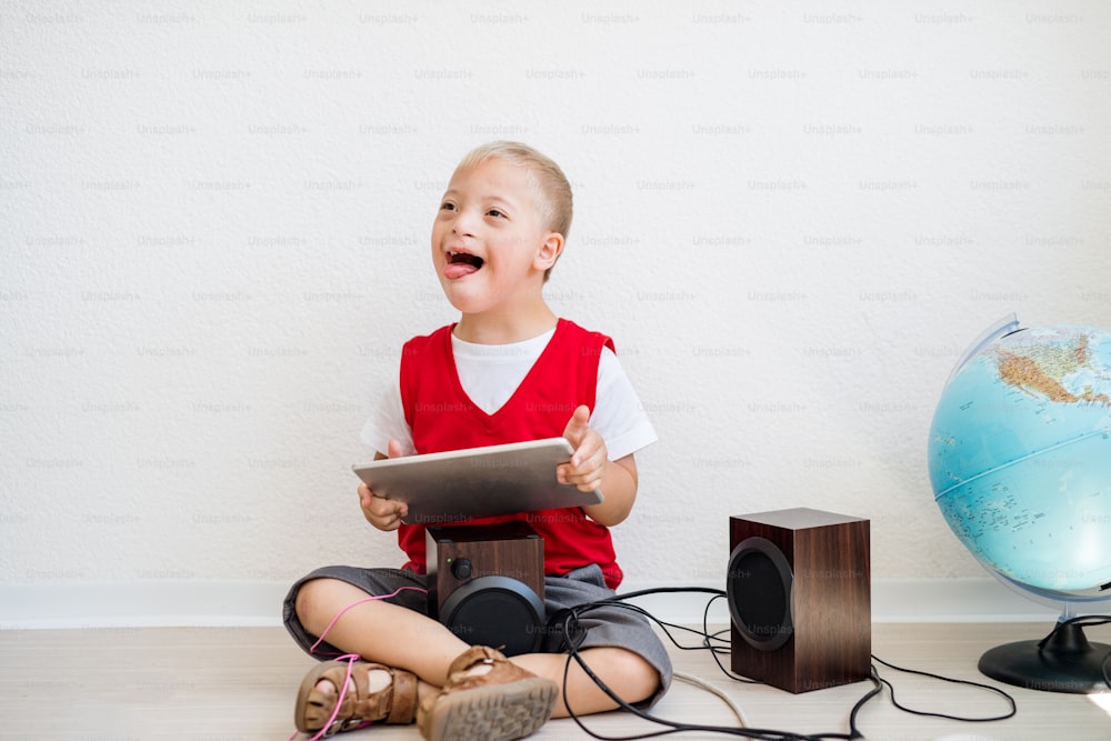 Um retrato de um menino feliz da escola com síndrome de Down com tablet sentado no chão, brincando com alto-falantes.
