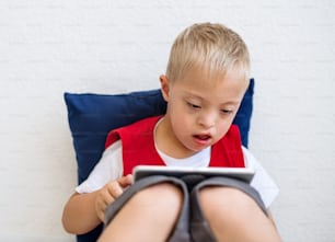 A portrait of happy down-syndrome school boy sitting on the floor, using tablet.