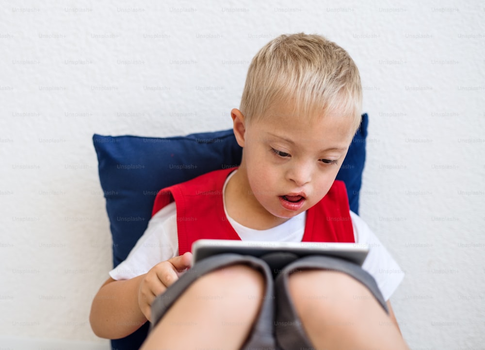 A portrait of happy down-syndrome school boy sitting on the floor, using tablet.
