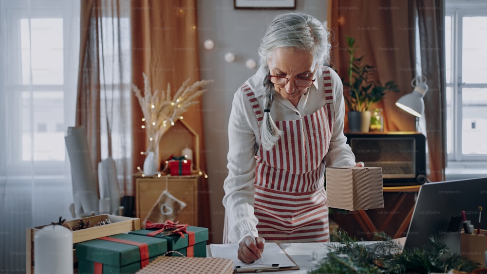 Una mujer mayor empacando regalos de Navidad y escribiendo notas para enumerar en el interior, concepto de pequeña empresa.