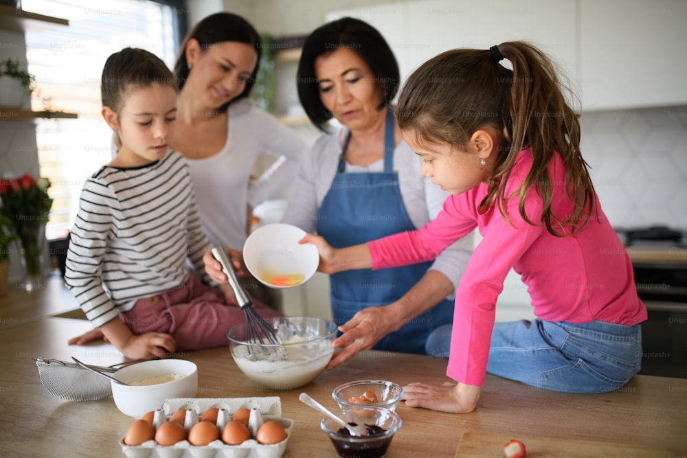 Meninas pequenas felizes com a mãe e uma avó fazendo mistura de panquecas dentro de casa, cozinhando.