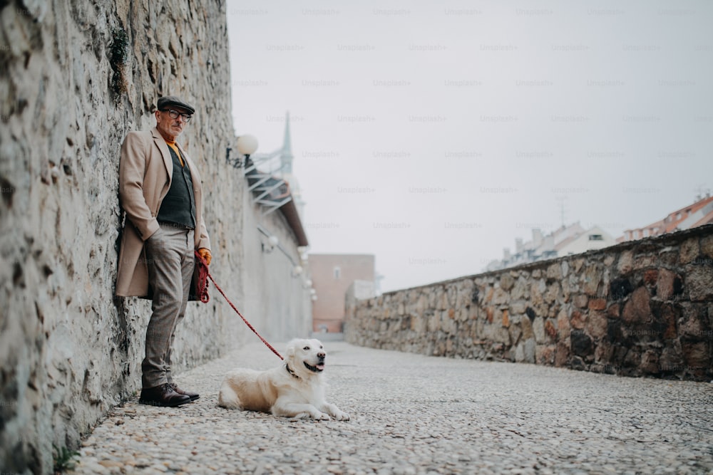A senior man looking at camera and leaning against the wall when walking his dog outdoors in city.