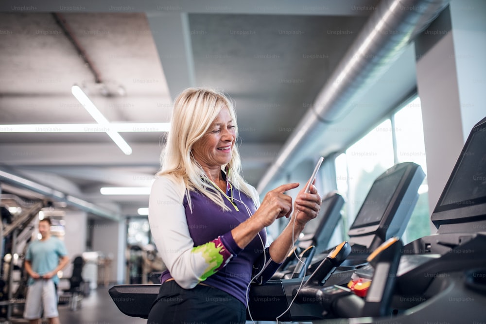 A happy senior woman with earphones and smartphone in gym doing cardio work out exercise.