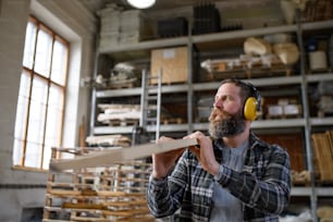 A mature male carpenter with hearing protectors carrying wooden board, indoors in carpentery workshop. Small business concept.