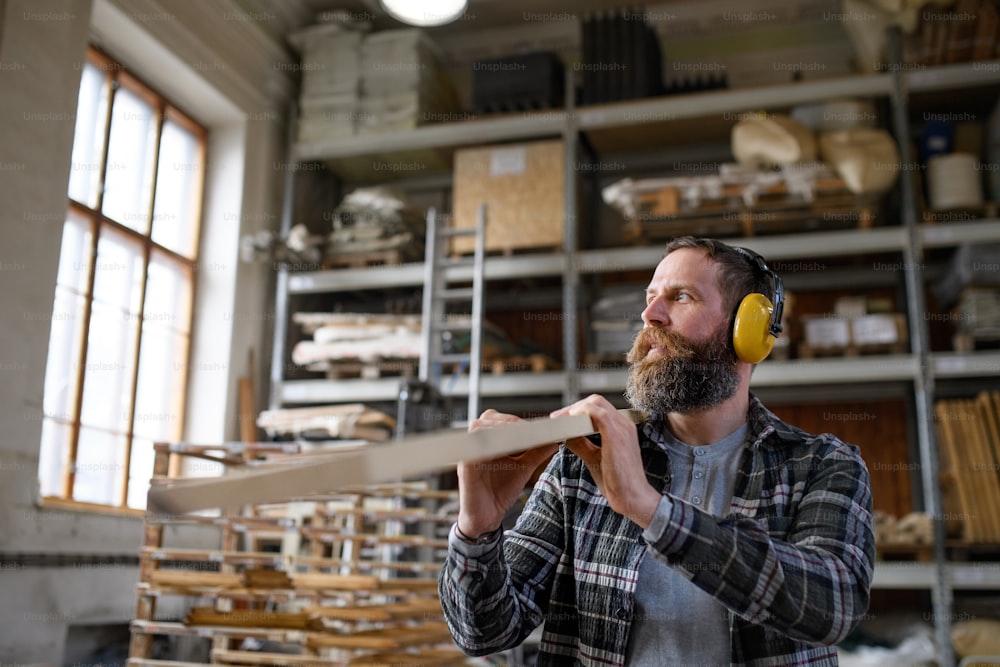 Un falegname maschio maturo con protezioni per l'udito che trasporta una tavola di legno, all'interno di un laboratorio di falegnameria. Concetto di piccola impresa.