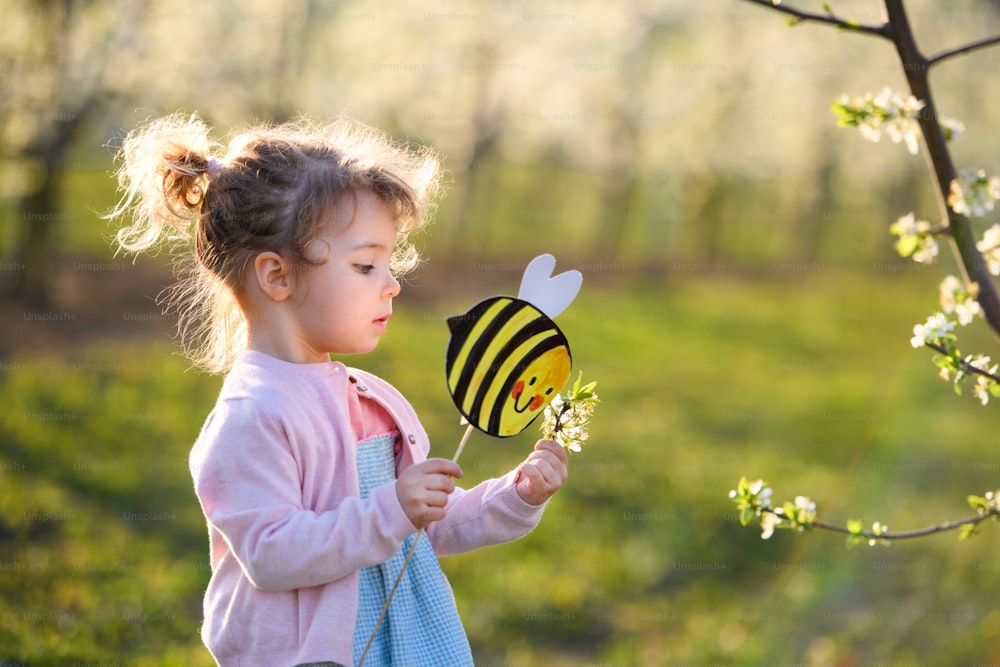 Seitenansicht eines kleinen Kleinkindes, das im Frühling draußen im Obstgarten steht und Papierbienen hält.