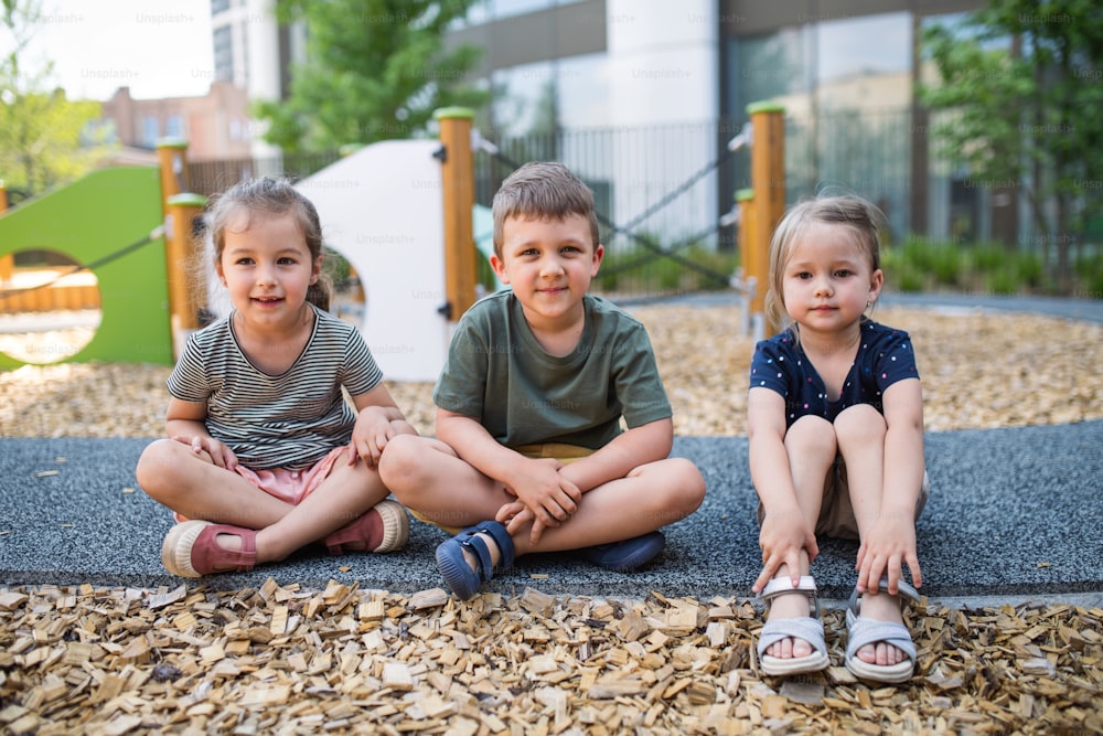 A group of small nursery school children sitting outdoors on playground, looking at camera.