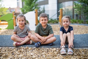 A group of small nursery school children sitting outdoors on playground, looking at camera.