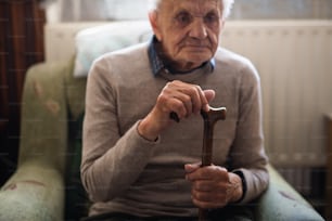 Un anciano triste con bastón sentado en un sillón en el interior de su casa, descansando.