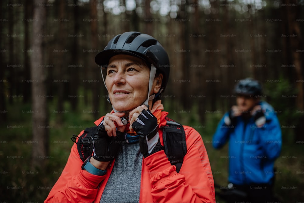 A senior couple bikers putting on cycling helmet outdoors in forest in autumn day.