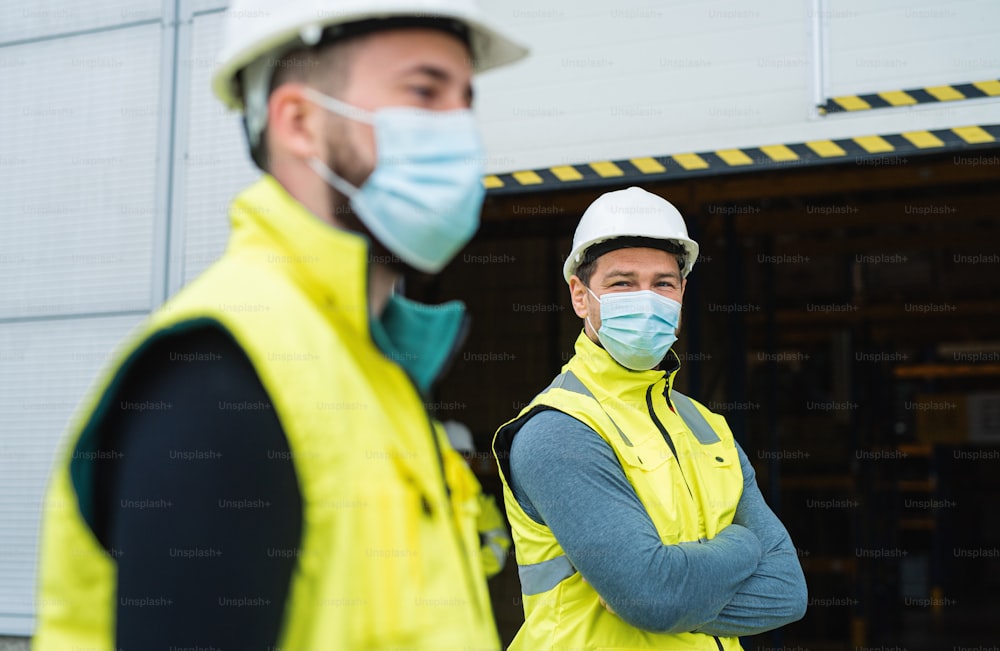창고 앞에 서 있는 안면 마스크를 쓴 두 남자 노동자, 코로나바이러스 개념.