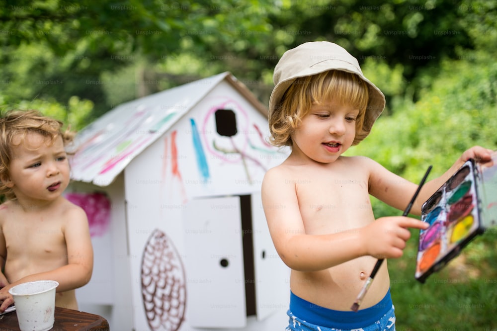 Portrait of topless small blond boy and girl with hat painting outdoors in summer.