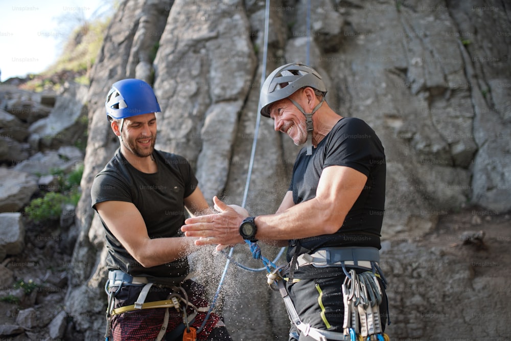 Portrait of senior man with instructor using chalk climbing rocks outdoors in nature, active lifestyle.