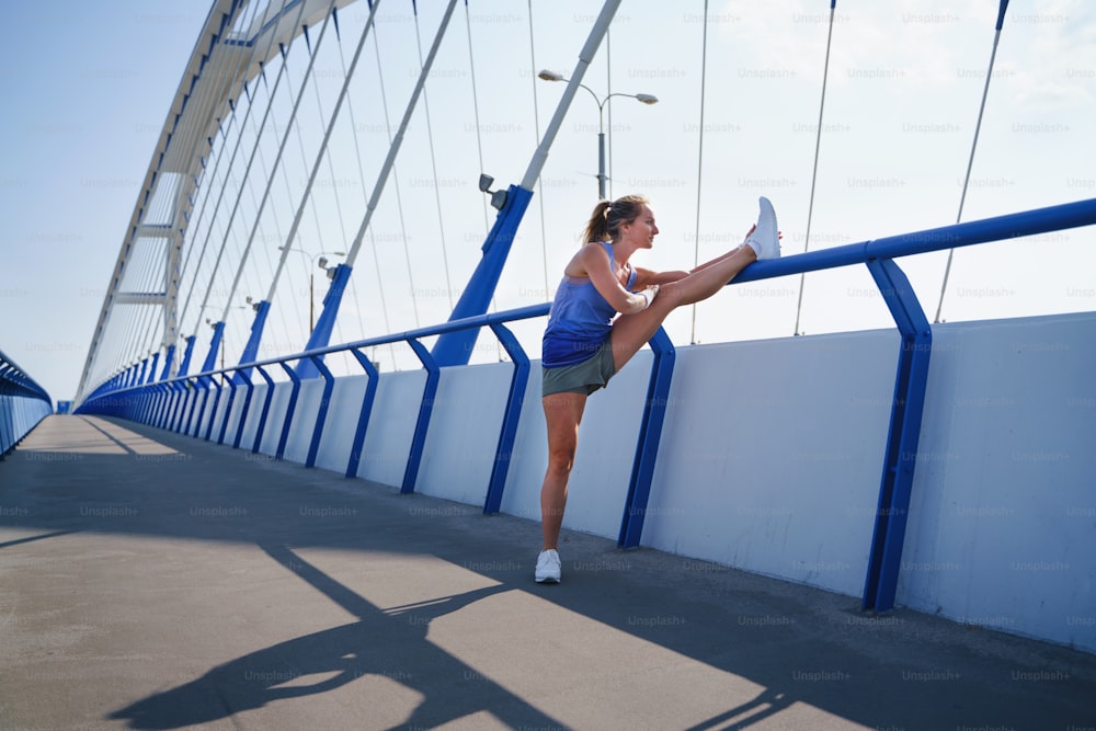 Una donna runner adulta media che si allunga all'aperto sul ponte in città, concetto di stile di vita sano.