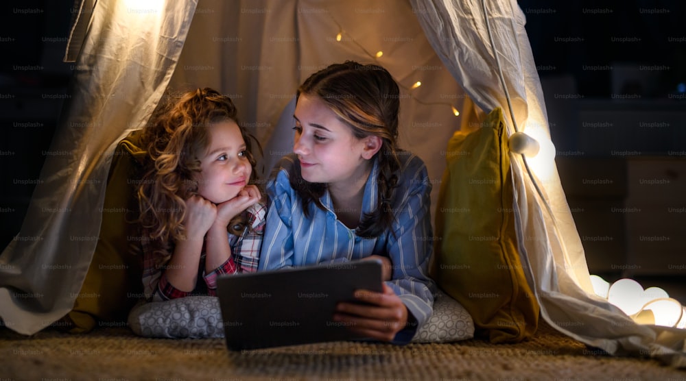 Sisters indoors at home, using tablet in pajamas in tent. Lockdown concept.