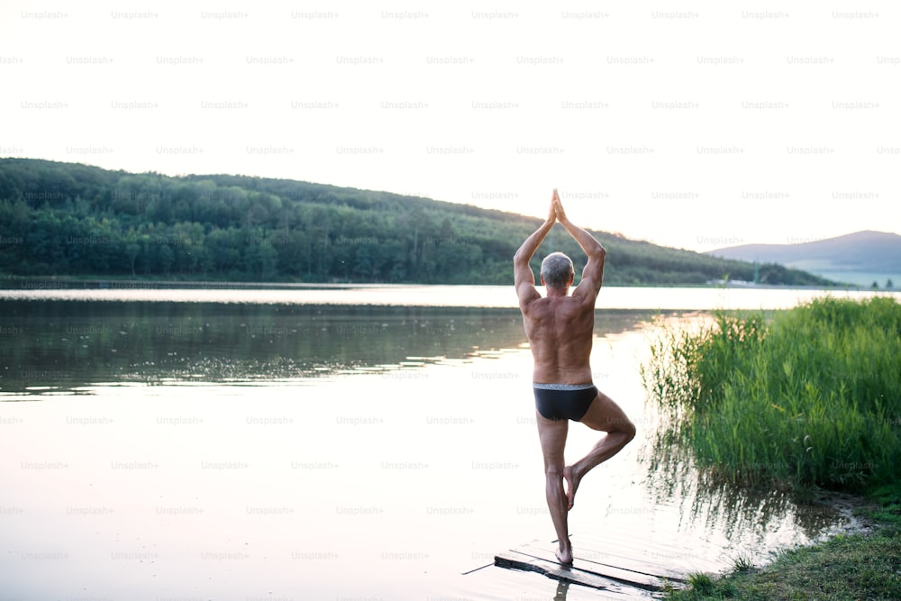 Vista traseira do homem sênior em traje de banho em pé junto ao lago ao ar livre fazendo yoga. Espaço de cópia.