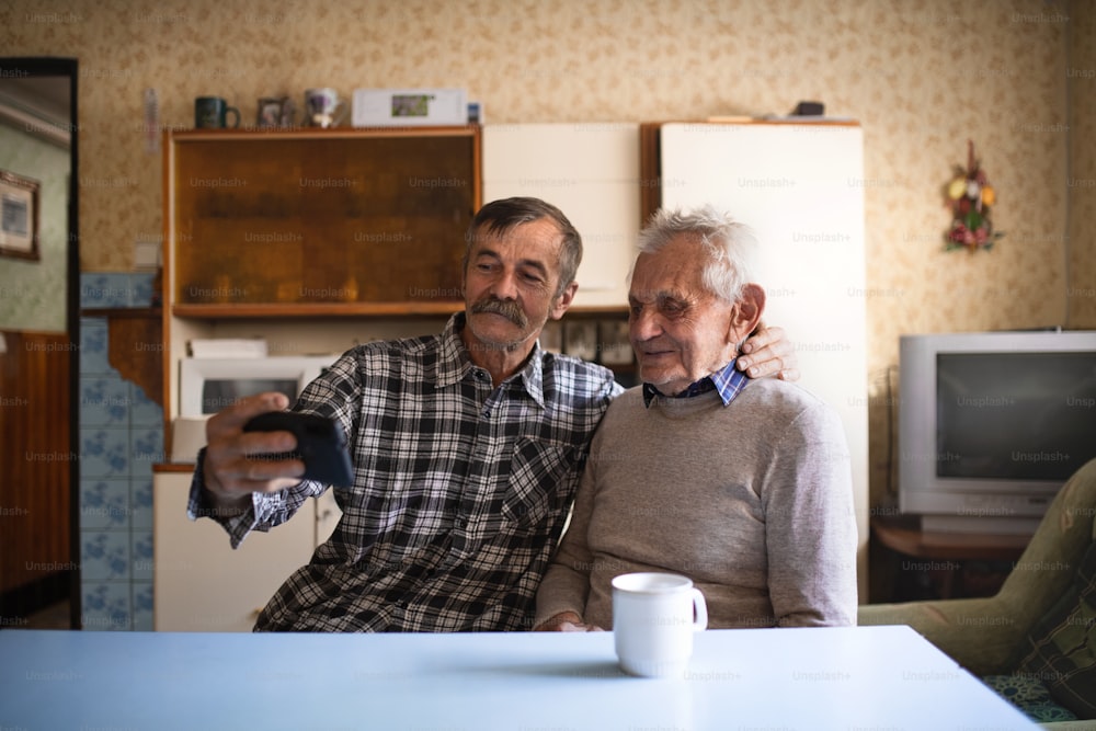 A portrait of man with elderly father sitting at the table indoors at home, taking selfie.