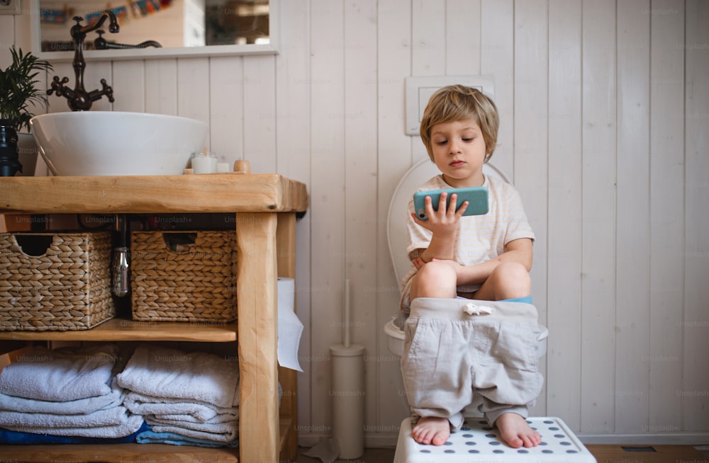 Un retrato de un lindo niño pequeño sentado en el inodoro en el interior de la casa, usando un teléfono inteligente.