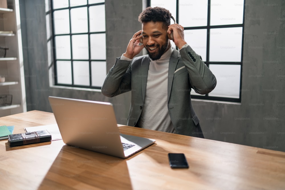 Joven hombre de negocios afroamericano feliz con un teléfono inteligente que trabaja en una computadora portátil en el interior de la oficina