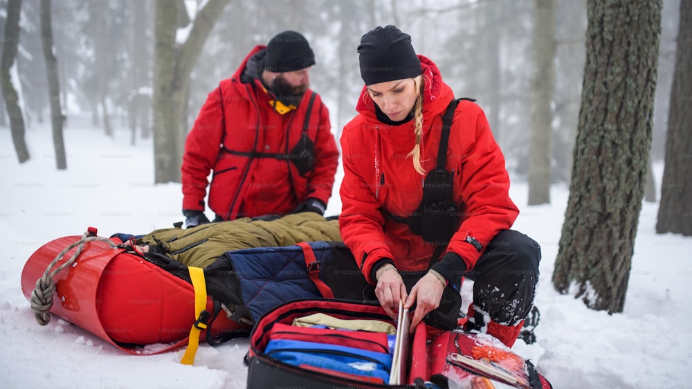 Paramedics from mountain rescue service provide operation outdoors in winter in forest, injured person in stretcher.