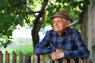 A portrait of elderly man standing outdoors in garden, leaning on wooden fence.