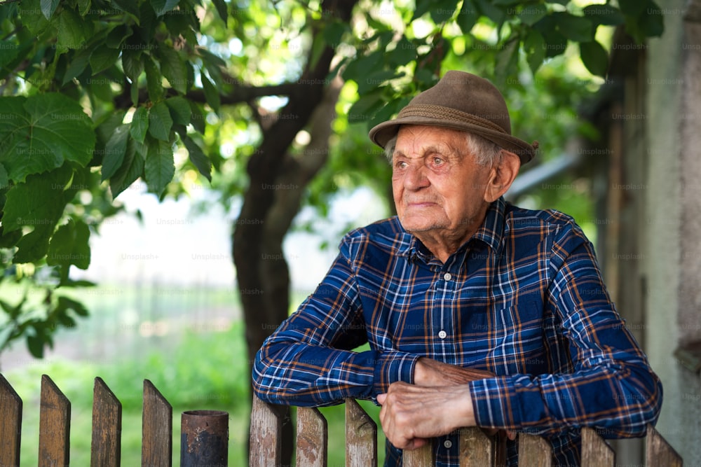 A portrait of elderly man standing outdoors in garden, leaning on wooden fence.