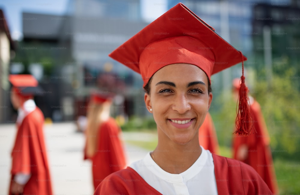 A portrait of cheerful university student with cap and gown looking at camera outdoors, graduation concept.