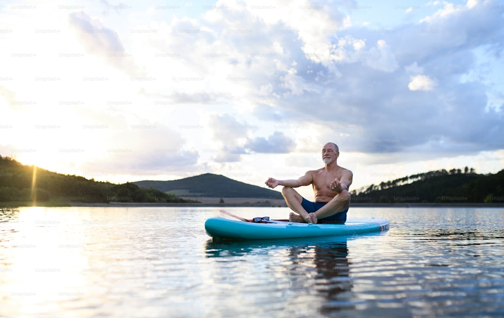 Senior man on paddleboard on lake in summer, doing yoga meditation exercise.
