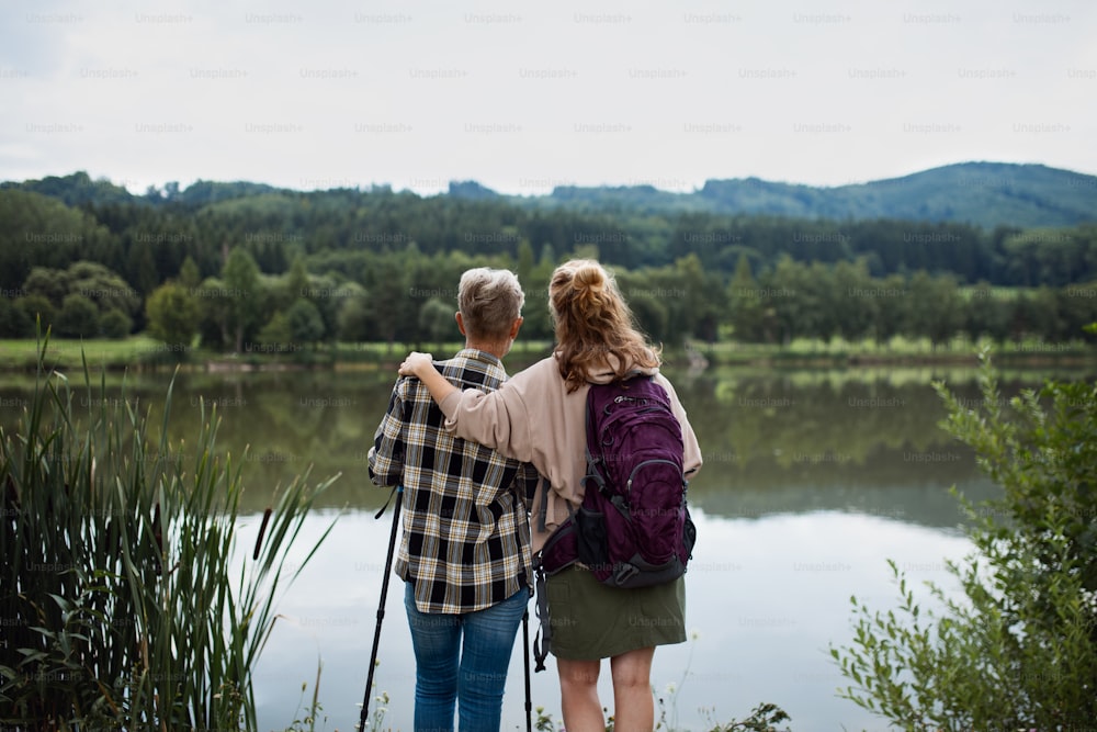 A rear view of senior mother embracing with adult daughter when standing by lake outdoors in nature