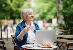 Portrait of senior woman with coffee sitting outdoors in cafe, using laptop.