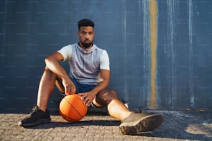 A young man with basketball doing exercise outdoors in city, sitting and resting.