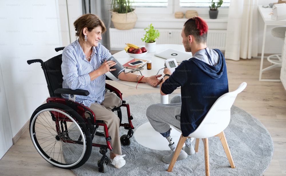 Two disabled people in wheelchair sitting at the table indoors at home, measuring blood pressure.