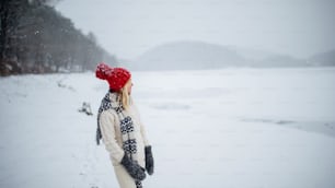 Happy senior woman with hat outdoors on a walk in snowy nature, standing. Copy space.