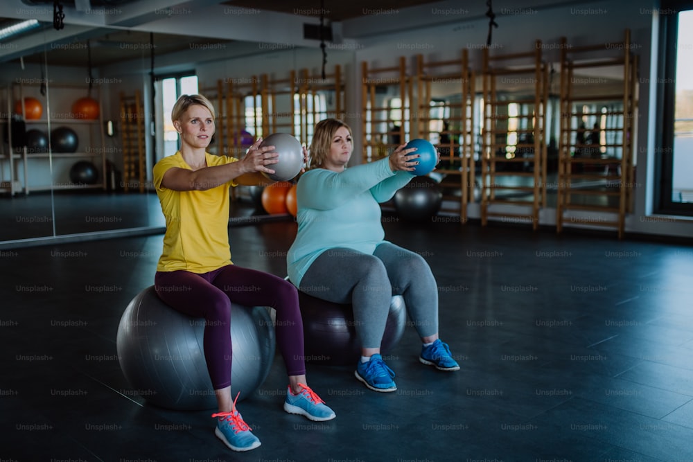 Una mujer con sobrepeso con mancuernas sentada en una pelota fintess y haciendo ejercicio con un entrenador personal en el gimnasio