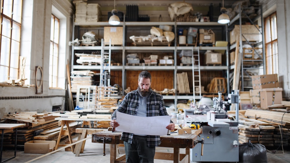 Un retrato de un carpintero masculino maduro mirando planos planos en un taller de carpintería. Concepto de pequeña empresa.