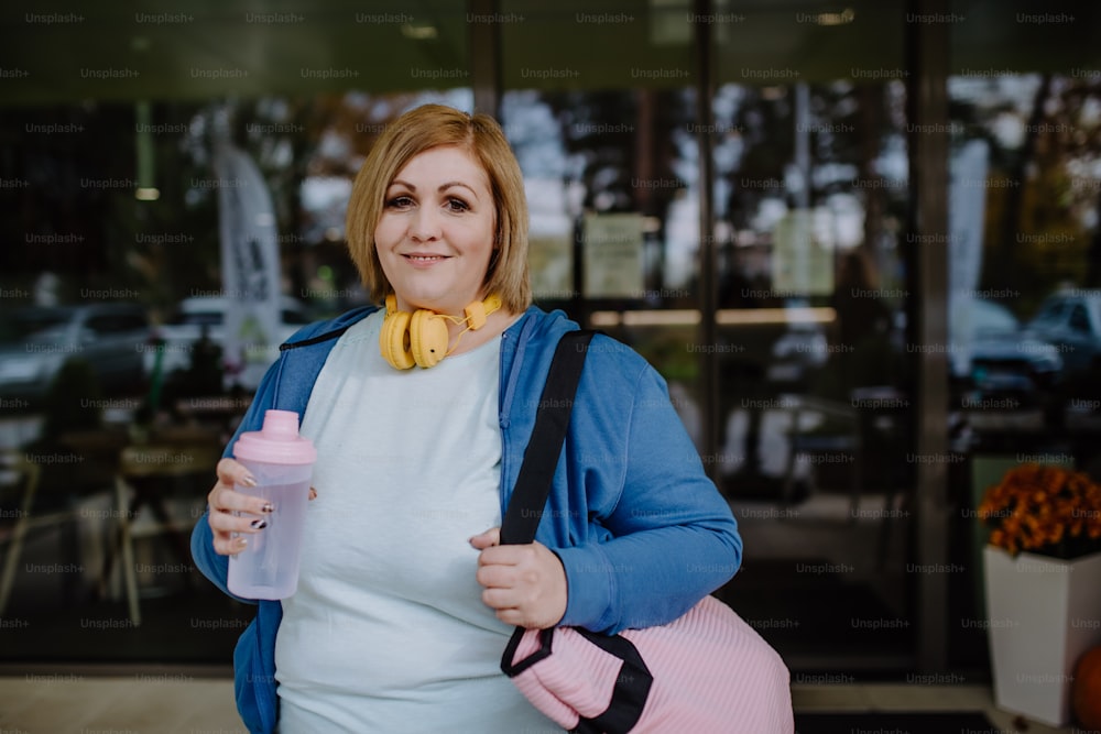 A happy overweight woman in sports clothes looking at camera outdoors in front of fitness center