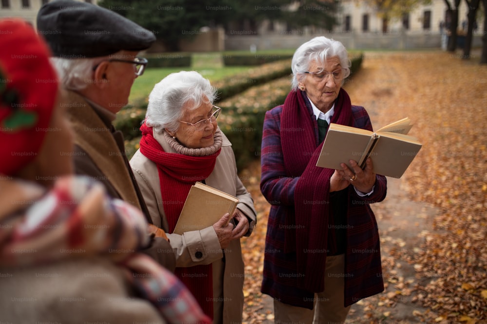 A group of happy senior friends with book on walk outdoors in park in autumn, reading and talking.