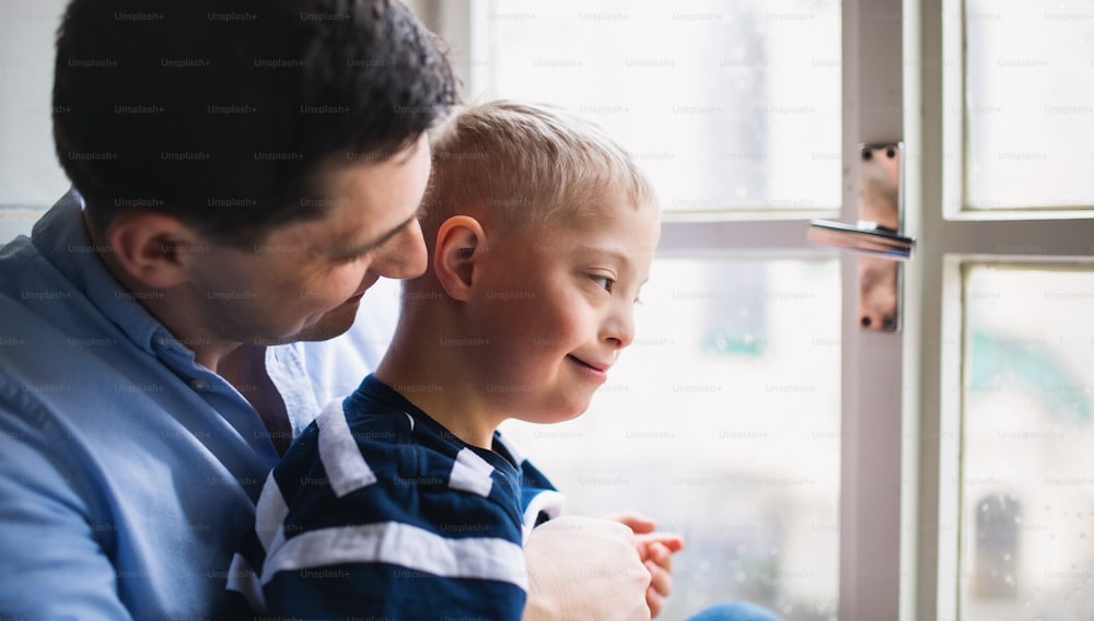 Père mûr avec un fils heureux du syndrome de Down à l’intérieur à la maison, regardant par la fenêtre.