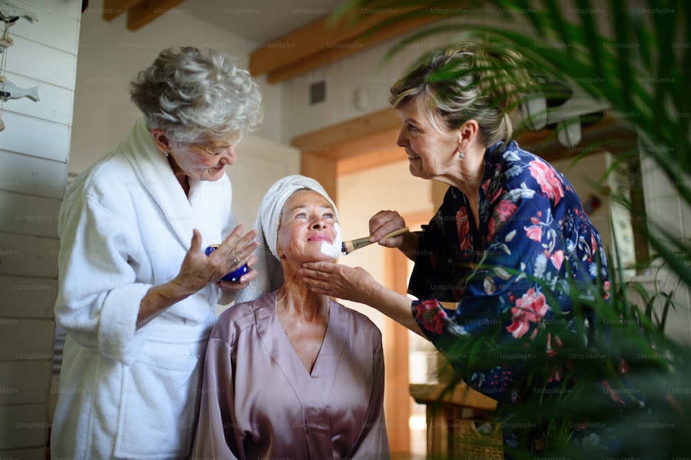 Happy senior women friends in bathrobes applying face mask indoors in a bathroom, selfcare concept.