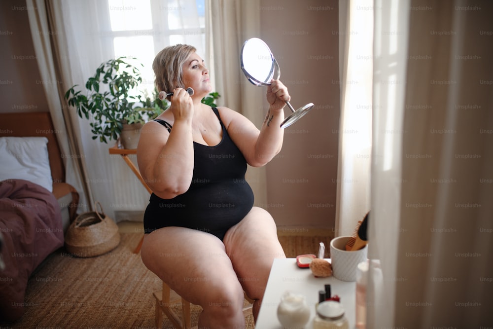 Uma mulher gorda sentada e olhando para o espelho e aplicando maquiagem em casa, conceito de autocuidado.