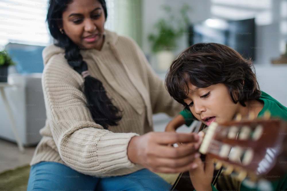 A little multiracial boy learning to play the guitar with his mother at home.