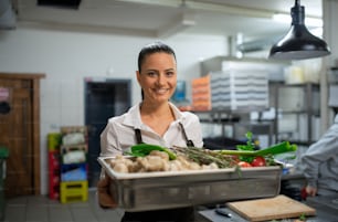 Una chef donna che trasporta un vassoio con verdure fresche e guarda la telecamera nella cucina del ristorante.