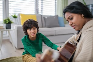 A little multiracial boy learning to play the guitar with his mother at home.