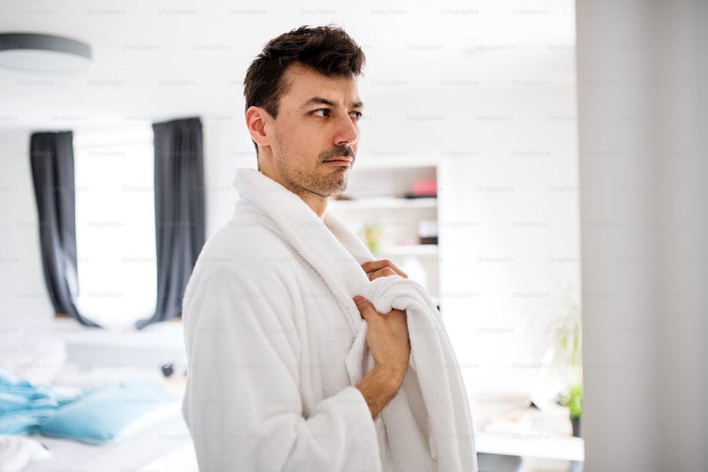 Young man with bathrobe in the bedroom, looking in the mirror. A morning routine.