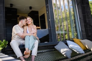 A young couple sitting and cuddling in hammock terrace in their new home in tiny house in woods, sustainable living concept.