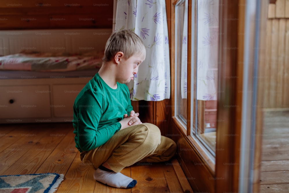 Um garotinho triste com síndrome de Down sentado no chão e olhando pela janela em casa.